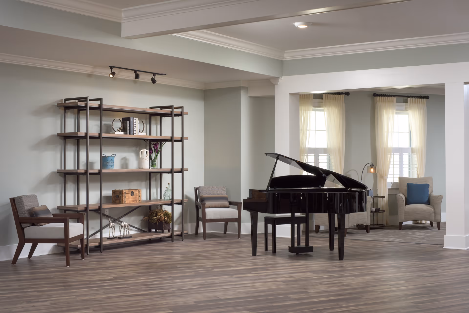 A spacious and well-lit living area featuring a black grand piano at the center, two armchairs with cushions, a tall open shelving unit with decorative items, and large windows with sheer curtains allowing natural light to fill the room.