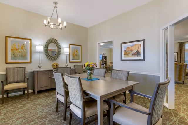 A dining room with a rectangular wooden table surrounded by six upholstered chairs. On the table is a vase with colorful flowers. The room has beige walls with white trim, two framed floral paintings, a round decorative mirror, and two wall-mounted lamps above a sideboard. A chandelier hangs from the ceiling, and doorways lead to other rooms.
