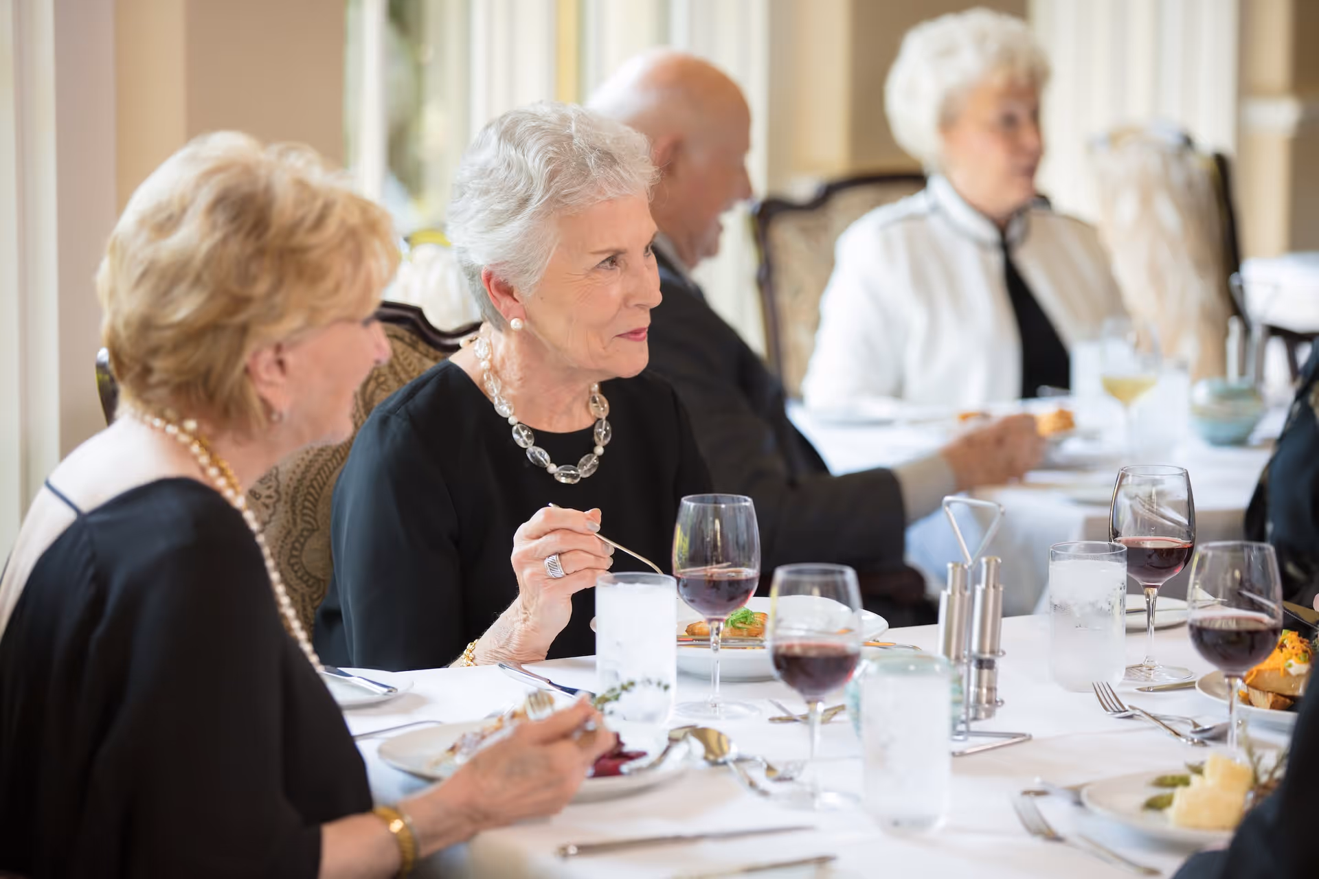 A group of elderly people sitting at a dining table in a well-lit room, enjoying a meal together with glasses of water and red wine on the table.