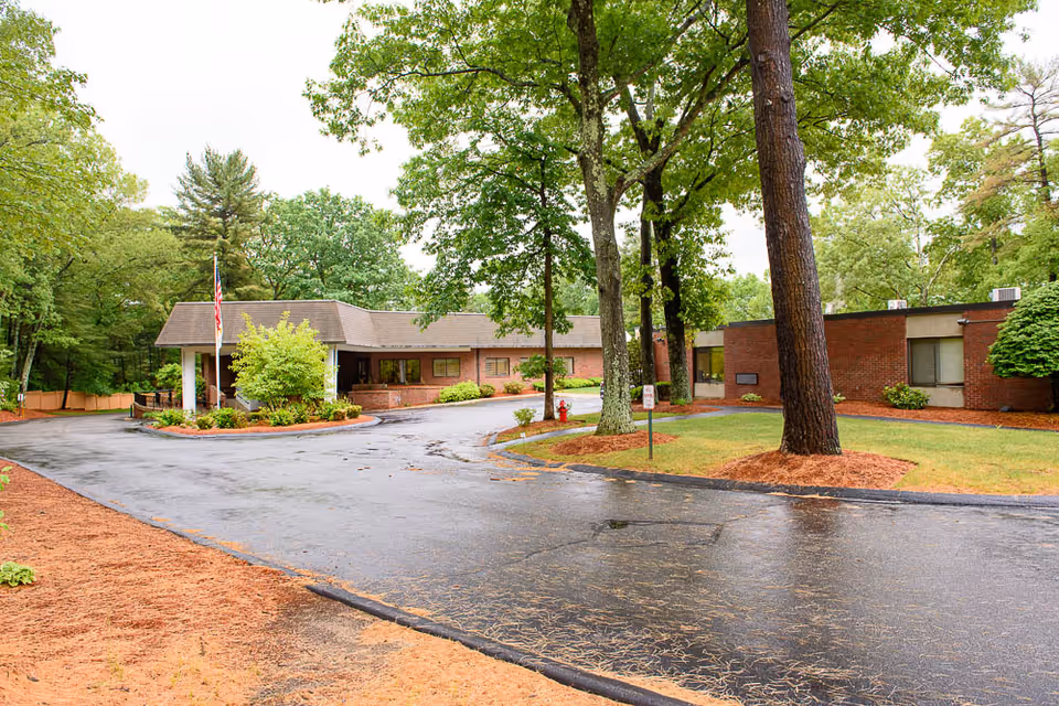 Front driveway and entrance of a single-story brick senior living facility surrounded by trees and landscaping.