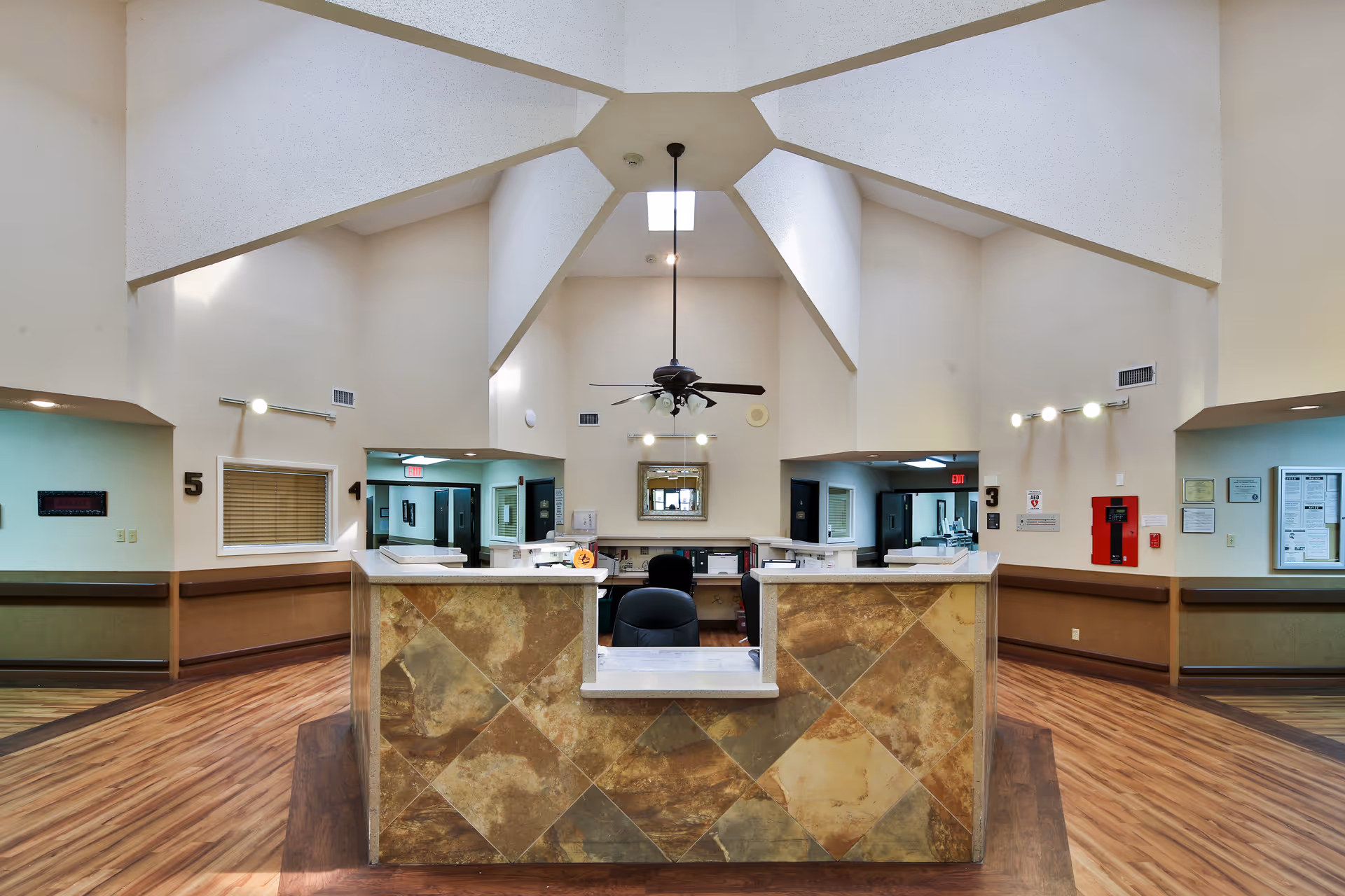 Interior view of a nursing and rehabilitation center reception area with a central desk featuring a tiled front. The ceiling has a unique geometric design with a ceiling fan and light fixture hanging down. There are numbered signs on the walls indicating different rooms or sections, and the floor has wood-like flooring.