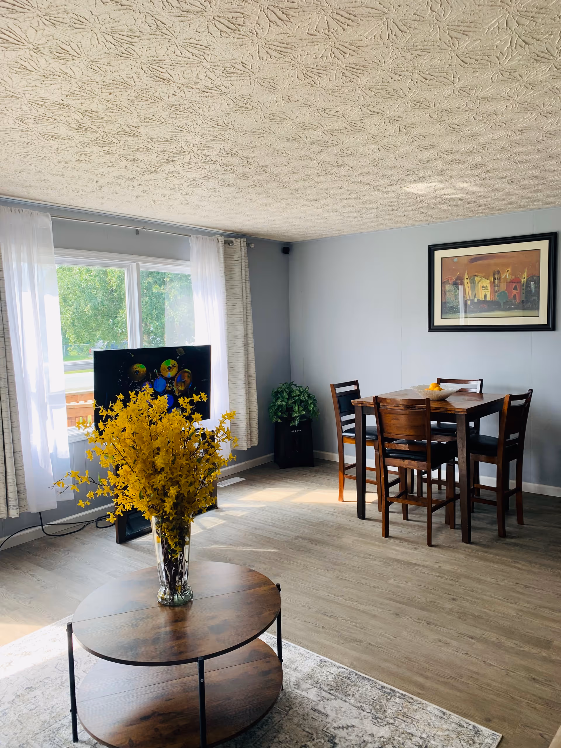 A bright living and dining area with light gray walls and a textured white ceiling. There is a round wooden coffee table with a glass vase filled with yellow flowers in the foreground. In the background, a wooden dining table with four chairs is set against the wall, which has a framed colorful painting. A flat-screen TV is positioned near a window with white curtains, and a green potted plant is placed in the corner. The floor is light wood, and sunlight streams in through the window.