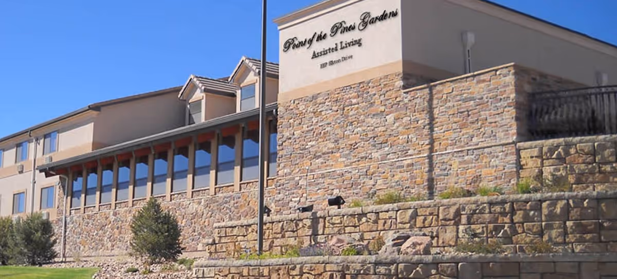 Exterior view of a stone-front assisted living building with a sign reading 'Point of the Pines Gardens' and terraced landscaping under a clear blue sky.
