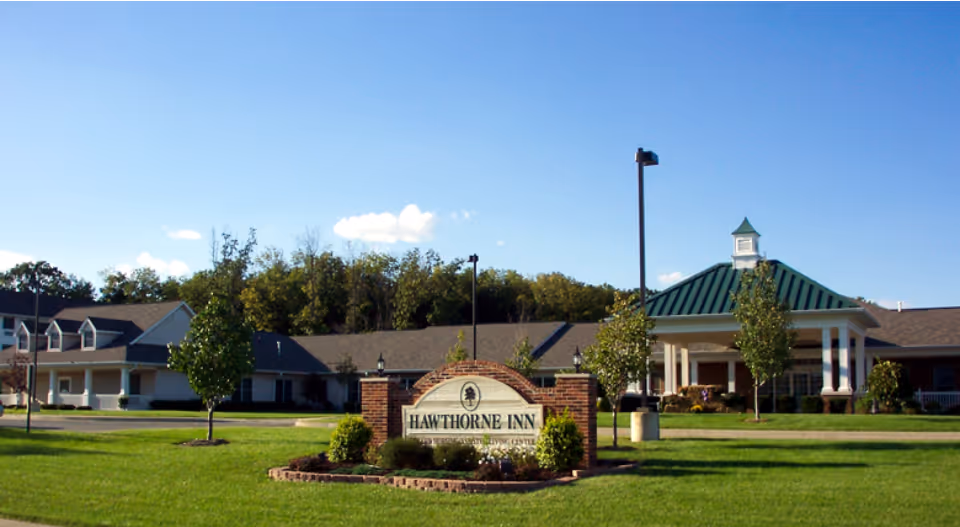 Exterior view of a senior living facility named Hawthorne Inn with a large green lawn, trees, and a clear blue sky. The building has a covered entrance with white columns and a green roof.