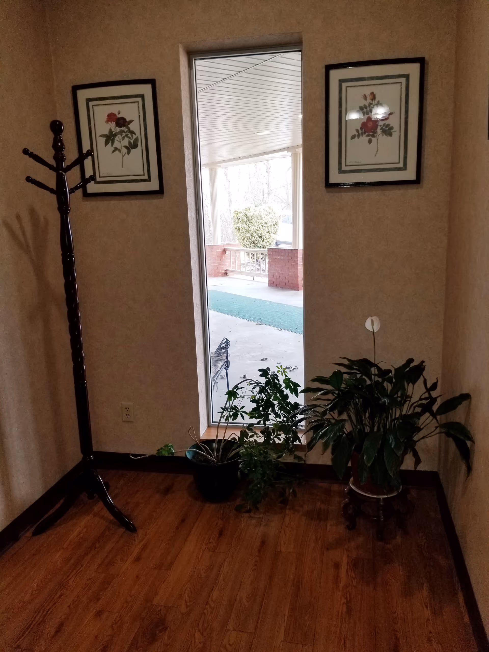A corner of a room with wooden flooring and beige walls. There is a tall wooden coat rack on the left side and two framed floral pictures hanging on the walls. Below the window, there are two potted plants, one on the floor and one on a small wooden stand. The window shows an outdoor covered area with a green mat and a brick railing.