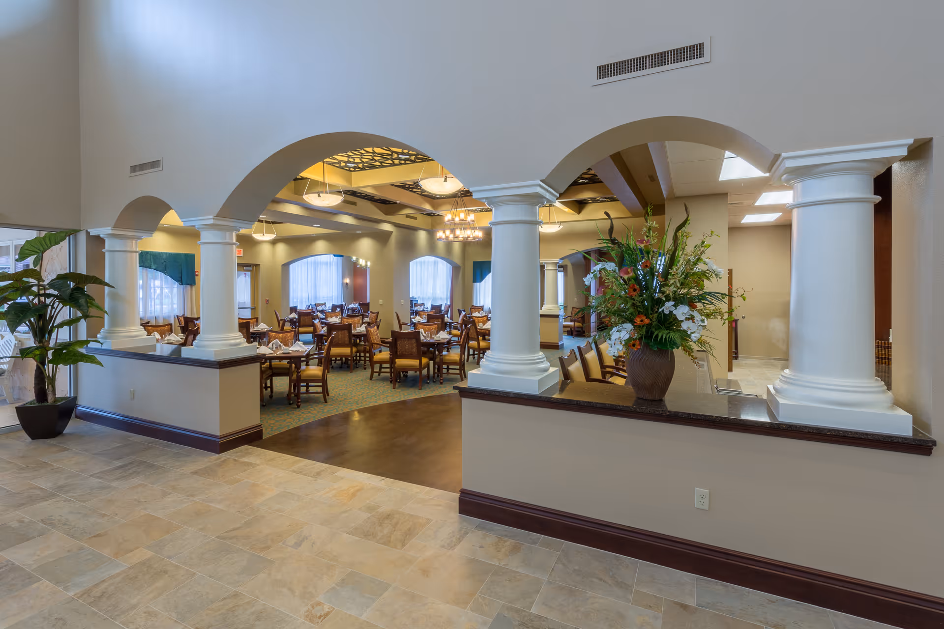 Spacious dining room with arched columns, multiple set tables and chairs, and a large floral arrangement by the entrance.