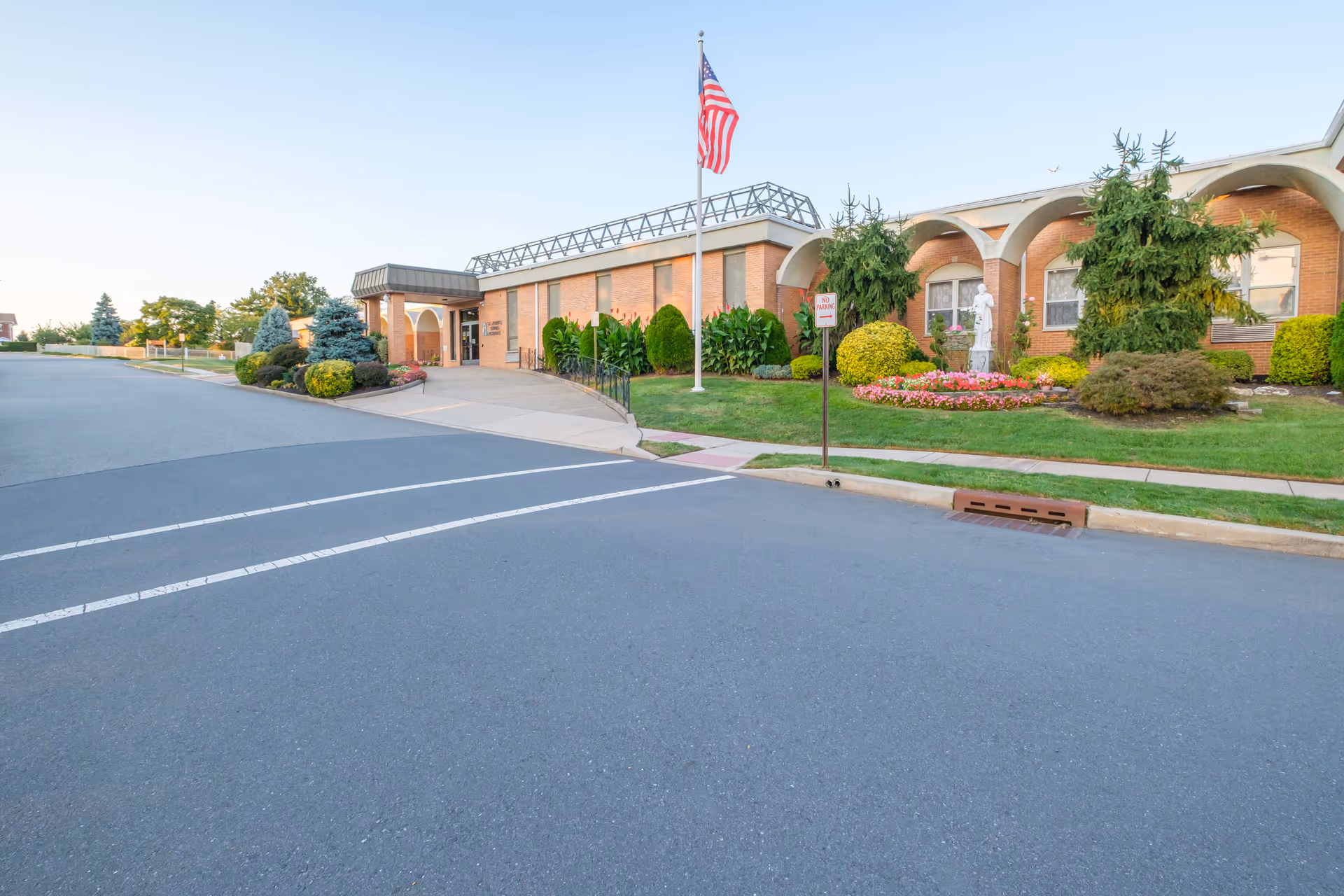 Exterior view of St Joseph's Senior Home - Assisted Living and Nursing Center showing a single-story brick building with arched windows, a landscaped garden with shrubs and flowers, an American flag on a flagpole, and a paved driveway with a no parking sign.
