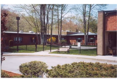 A view of the exterior of The Friendly Home Inc, featuring a landscaped area with trees, a bench, and a fenced yard.