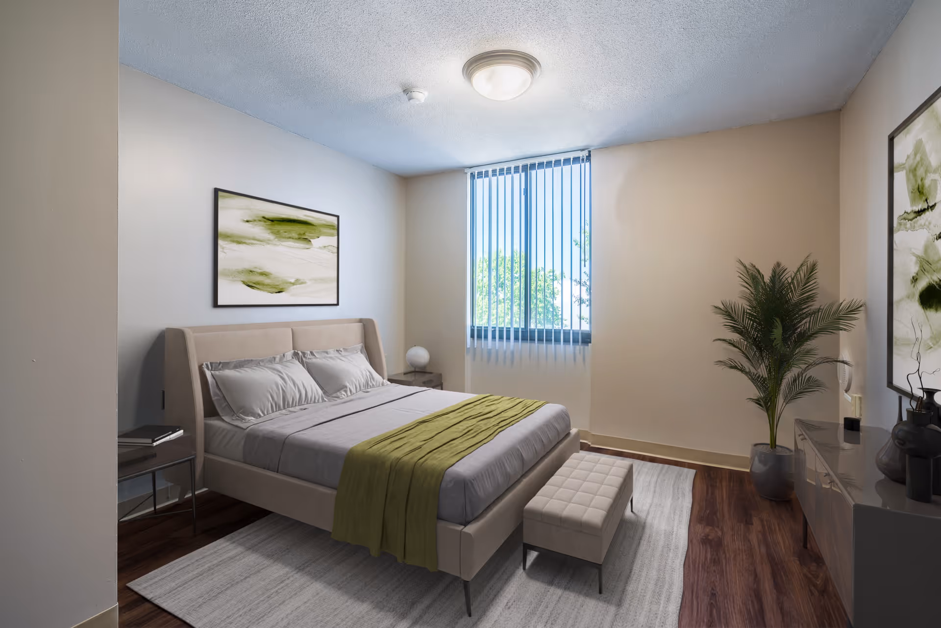 A neatly arranged bedroom with a beige upholstered bed featuring gray bedding and a green throw blanket. There is a small bench at the foot of the bed, a nightstand with a lamp, a large window with vertical blinds, a potted plant, and abstract green and white artwork on the walls. The floor is wooden with a light-colored area rug.
