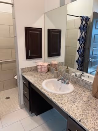 A bathroom with a granite countertop featuring a white sink and silver faucet. Above the sink is a large mirror reflecting a shower area with beige tiles and a blue patterned shower curtain. There are two dark wooden cabinets mounted on the wall above the countertop, and a small basket with toiletries is placed on the counter.