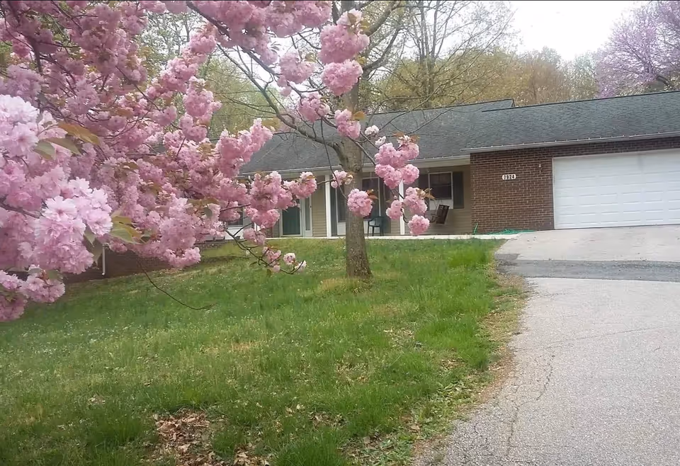 Front exterior view of a single-story brick and siding building with a green roof, a white garage door, and a porch with a swing and chairs. In the foreground, there is a grassy lawn and a tree with pink blossoms.