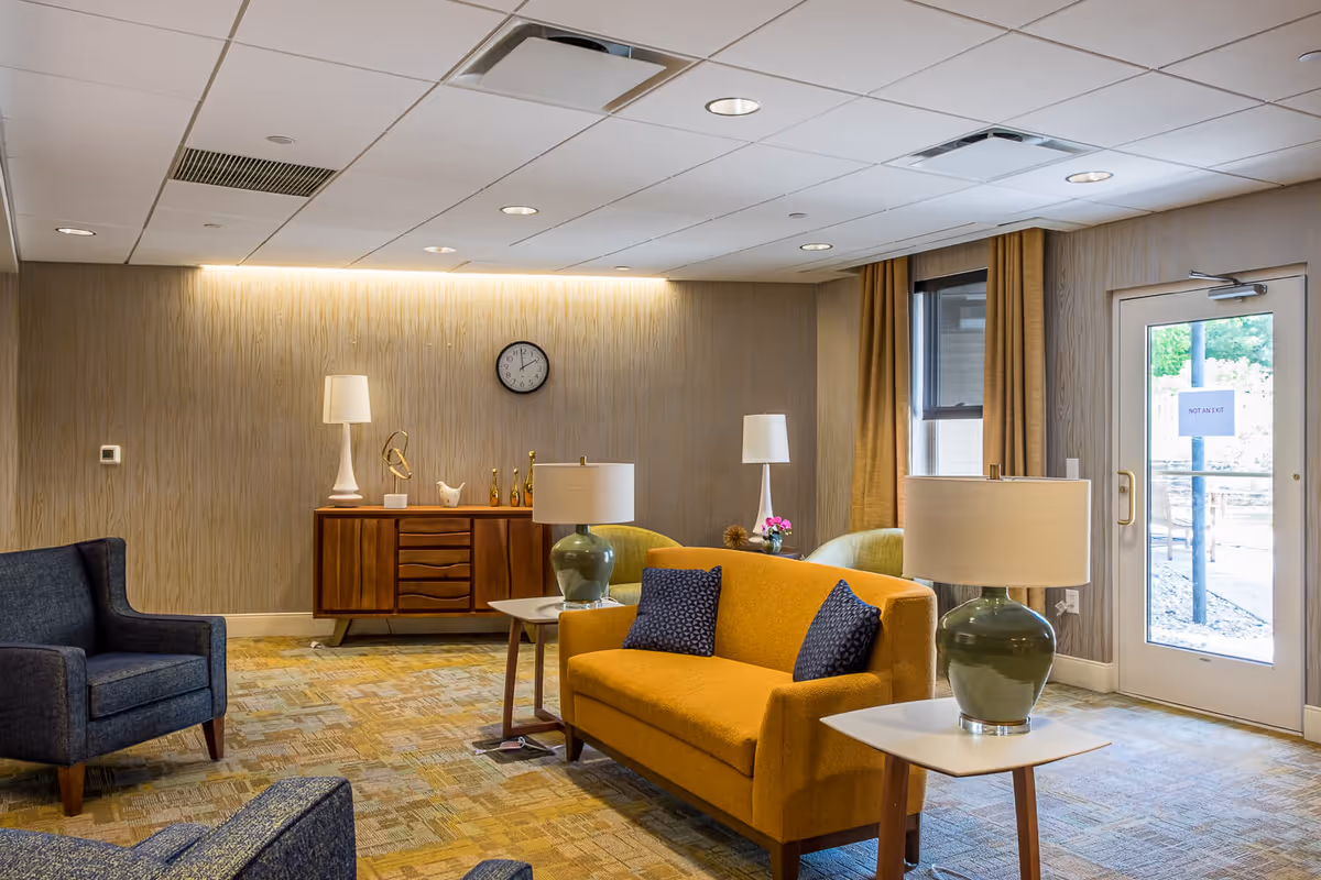 A bright senior living lounge with a mustard sofa, blue armchairs, side tables with lamps, a wooden sideboard and a door to the outside.