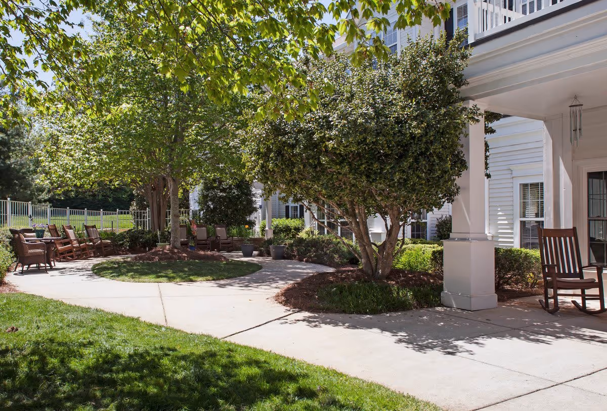 Shaded outdoor courtyard with a circular walkway, trees, potted plants and wooden chairs beside a white building.