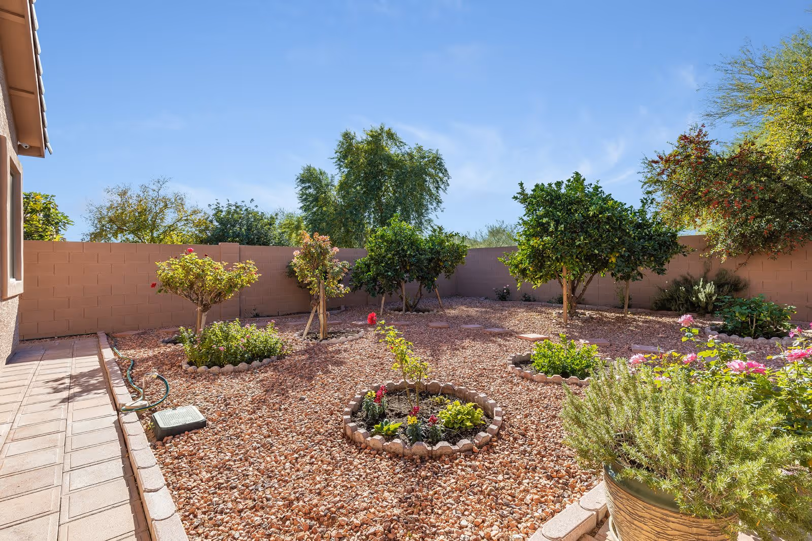 A sunny outdoor garden area with small trees and flower beds surrounded by a low brick wall. The ground is covered with reddish gravel, and there is a paved walkway on the left side. Various green plants and blooming flowers are visible under a clear blue sky.