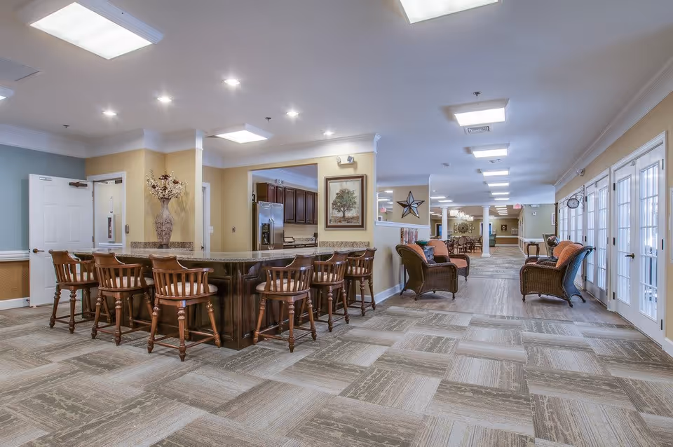 Spacious interior of a senior living facility featuring a large kitchen island with wooden bar stools, a seating area with wicker chairs and cushions, and multiple glass doors along one wall allowing natural light to enter. The space is well-lit with ceiling lights and decorated with framed artwork and a large vase with flowers.