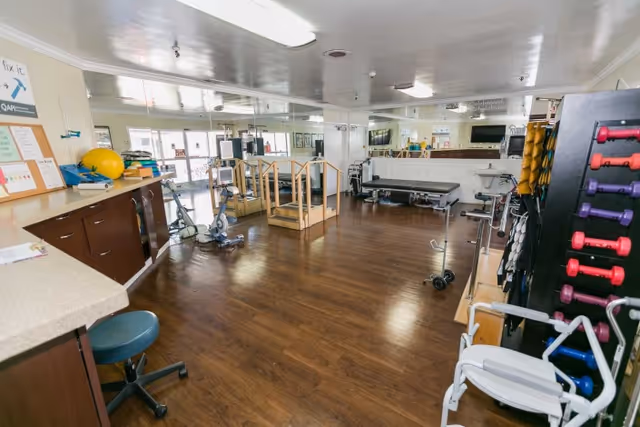 A spacious physical therapy or rehabilitation room with wooden flooring, exercise equipment including hand weights, therapy steps, stationary bikes, and a treatment table. There is a counter with a stool on the left side and large mirrors on the far wall reflecting the room.