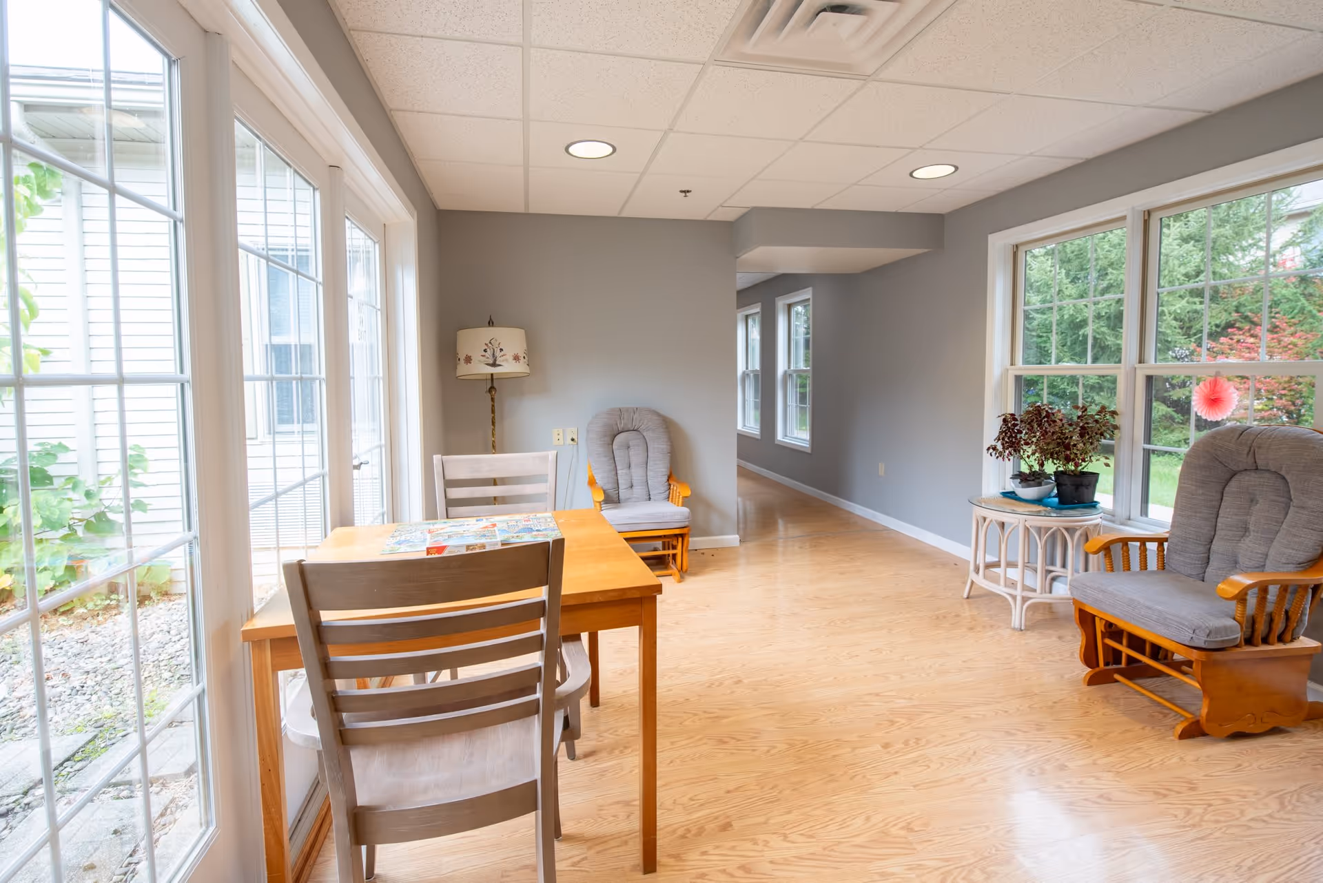 A bright room with large windows showing a garden outside. Inside, there is a wooden table with two chairs, a cushioned rocking chair, a small round table with potted plants, and a floor lamp with a floral lampshade. The room has light wood flooring and gray walls.