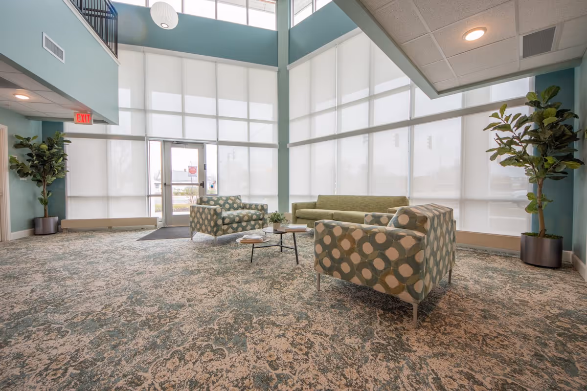 Bright senior living lobby with patterned armchairs, a green sofa, coffee table, large floor-to-ceiling windows and potted plants.