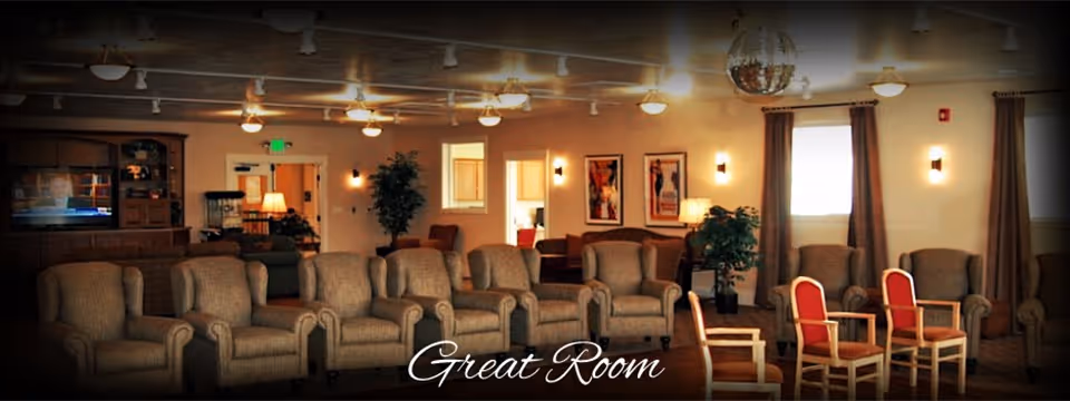 Communal great room with rows of upholstered armchairs, additional wooden chairs, a TV, lamps and plants in a senior living facility.