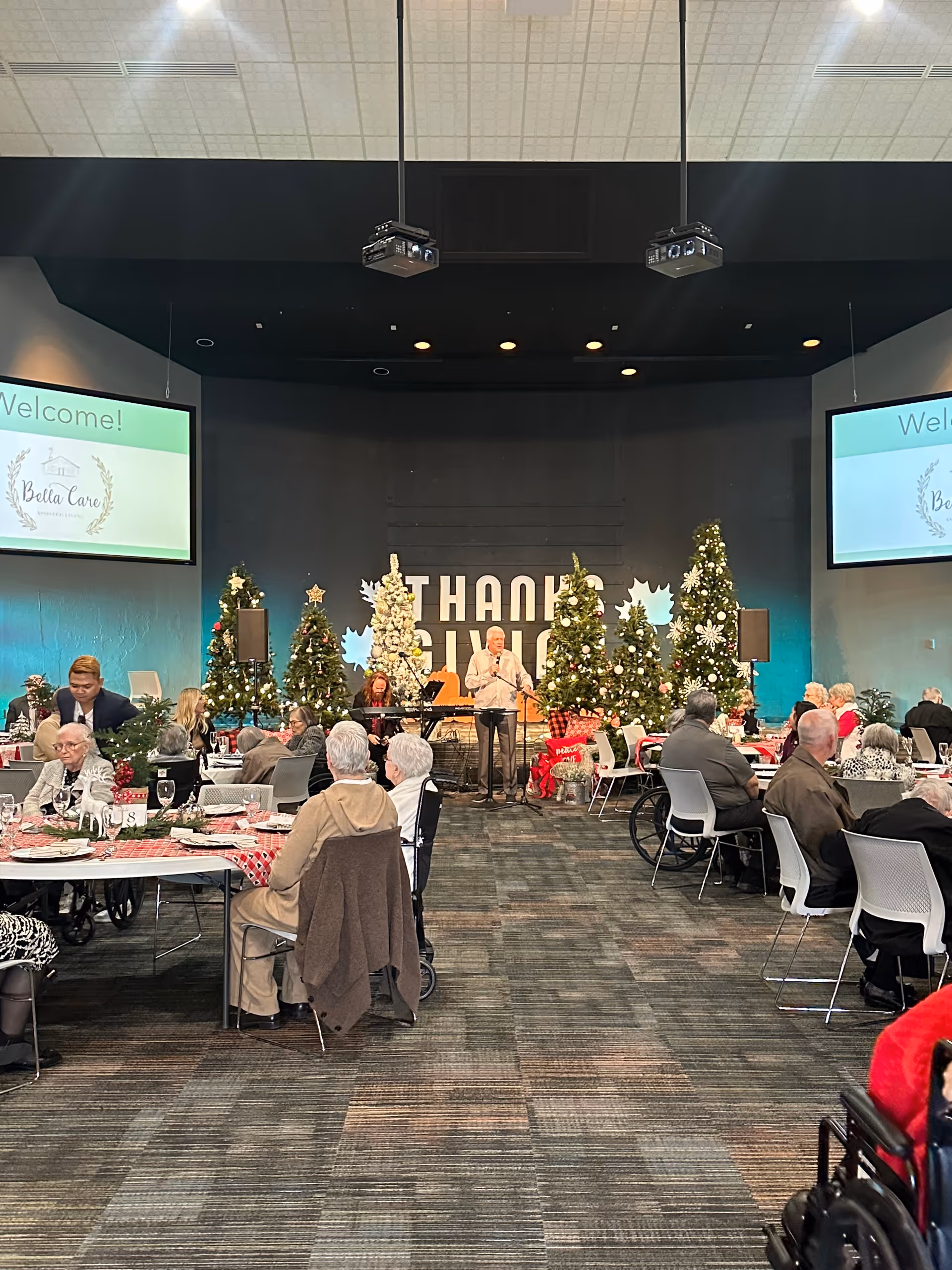 A large room decorated for a holiday event with several Christmas trees and festive decorations. Elderly people are seated at tables covered with red and white tablecloths, engaging in conversation. A man stands at a podium in front of a backdrop with the words 'THANK YOU' displayed. Two large screens on either side of the stage display a welcome message from Bella Care Home.