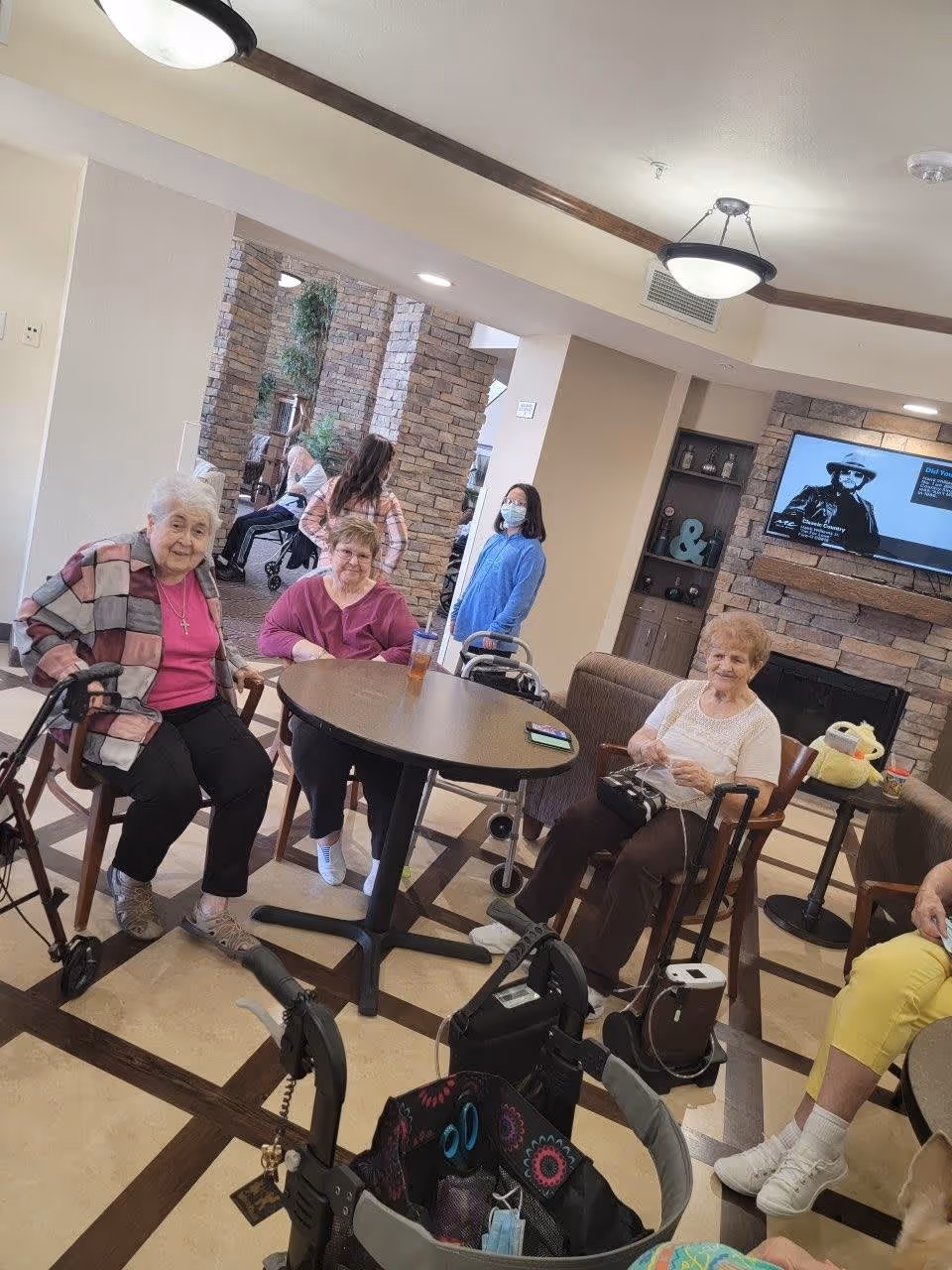 Several elderly residents sit around a table in a senior living facility common area with walkers, a TV, and a stone fireplace.