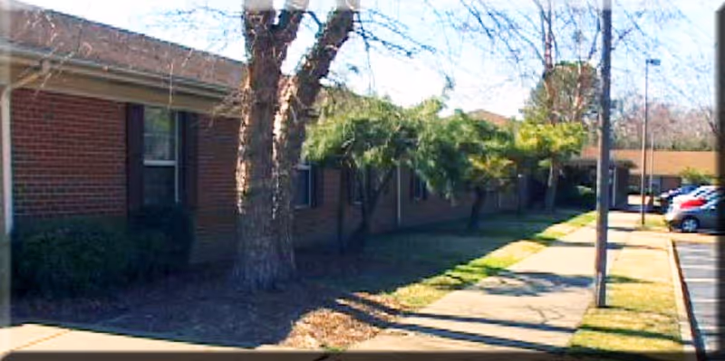 Single-story brick assisted living building front with a sidewalk, trees, and a parking area with cars.