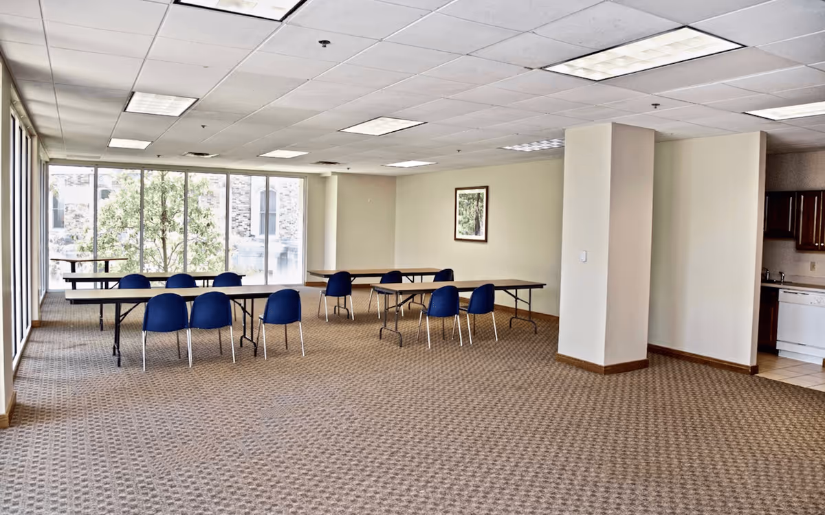 A spacious room with large windows letting in natural light, featuring several rectangular tables with blue chairs arranged around them. The room has beige walls, a patterned carpet, and a small kitchenette area with wooden cabinets and a dishwasher in the corner.