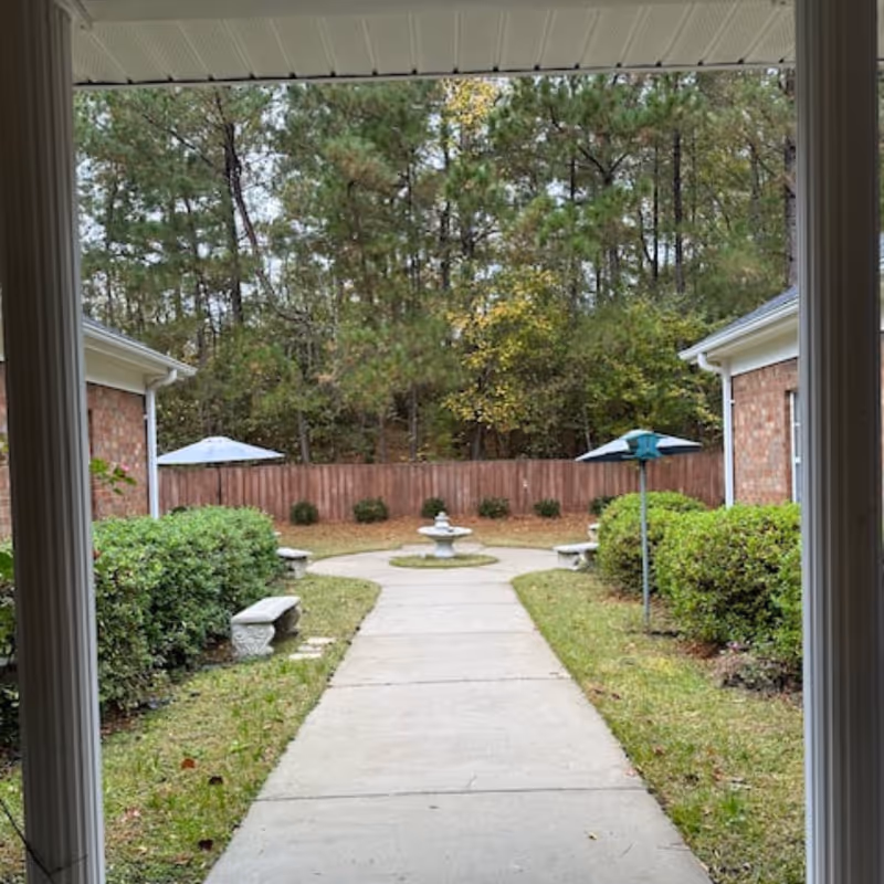 View of an outdoor courtyard area with a concrete pathway leading to a small fountain in the center. The courtyard is surrounded by bushes, two brick buildings on either side, and a wooden fence with trees in the background. There are two umbrellas visible behind the bushes.