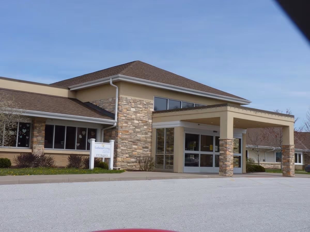 Exterior view of Lutheran Living facility showing the main entrance with a covered drop-off area, stone and beige siding, and a sign indicating the entrance.