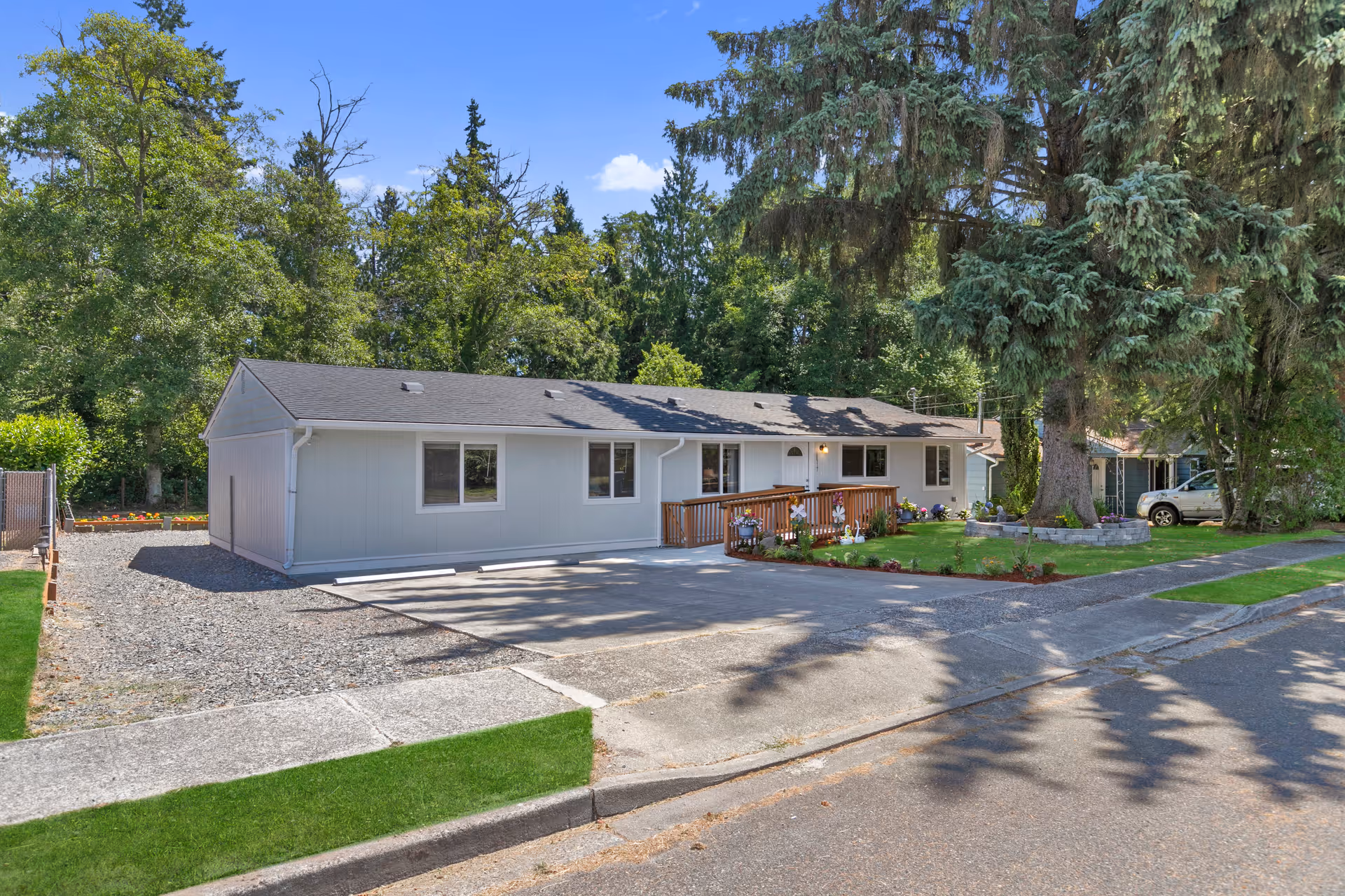 Single-story light gray building with a dark roof surrounded by trees and greenery. There is a wooden ramp leading to the entrance, a small landscaped garden with flowers, and a driveway in front. The scene is bright and sunny with a clear blue sky.