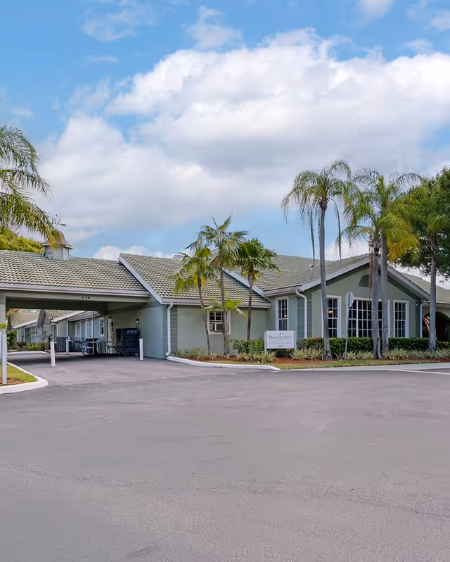 Exterior view of Brookdale Tequesta facility showing a single-story building with a covered entrance, palm trees, and a clear blue sky with some clouds.