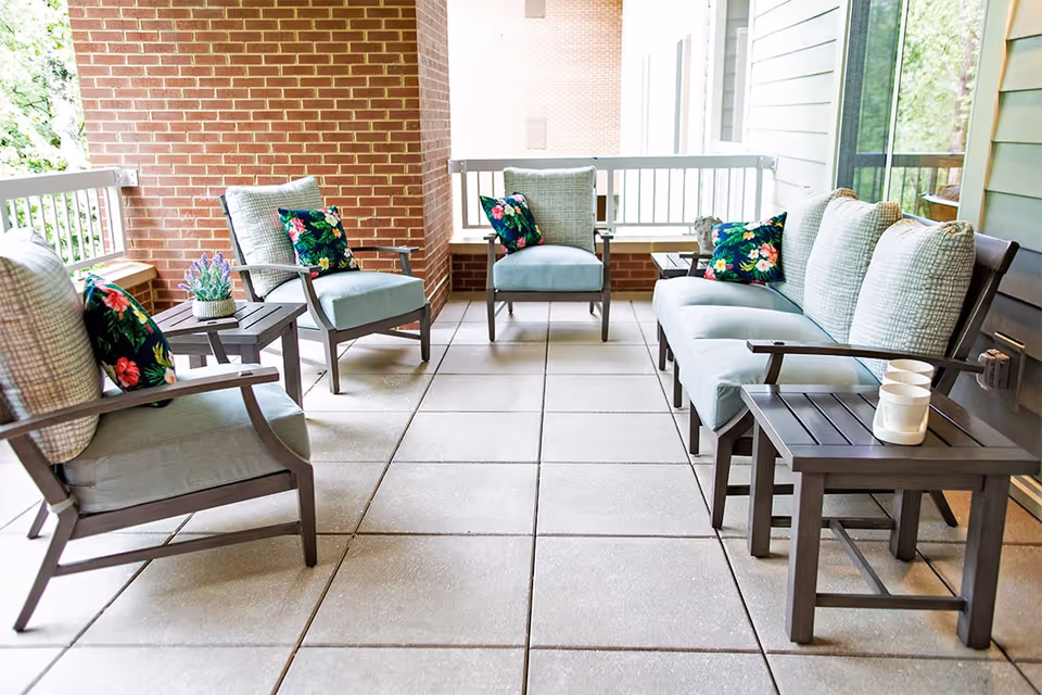 Outdoor patio area with cushioned seating including a sofa and chairs, decorated with floral patterned pillows and small side tables holding potted plants and cups.