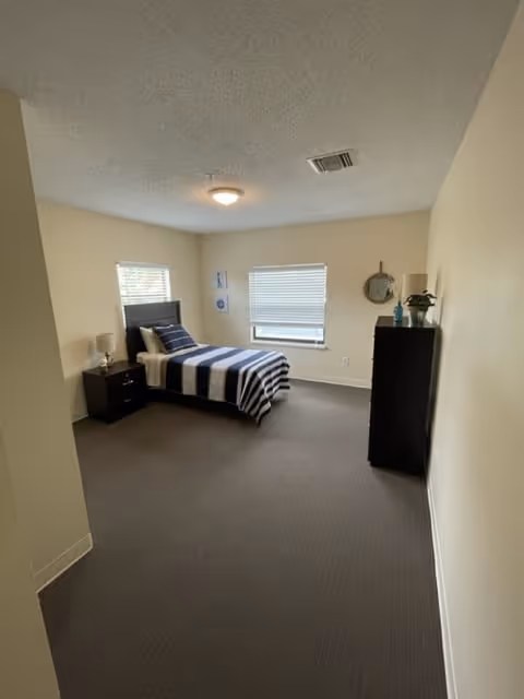 A simple bedroom with a single bed covered in a blue and white striped blanket, a dark wooden nightstand with a lamp, two windows with blinds, a dark wooden dresser with decorative items on top, and light beige walls and carpeted floor.