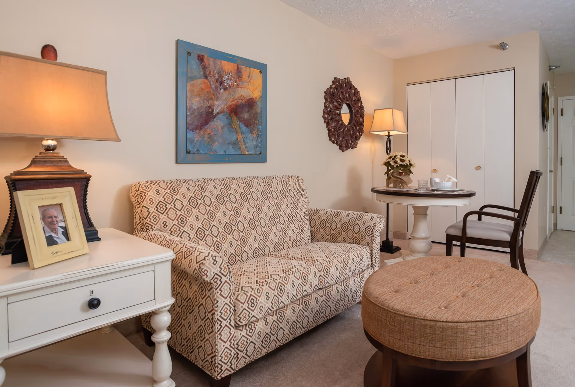 A cozy living room area featuring a patterned loveseat, a round cushioned ottoman, a small white side table with a lamp and a framed photo, a round table with a chair, a decorative wall mirror, and a colorful abstract painting on the wall.