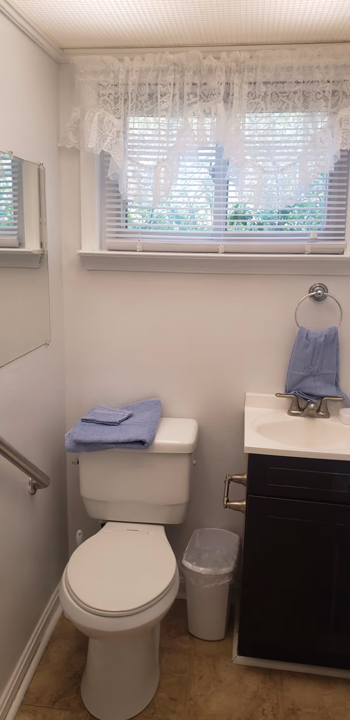 A small bathroom with a white toilet, a blue towel folded on top of the toilet tank, a white trash bin beside the toilet, a dark-colored vanity with a sink, a blue hand towel hanging on a ring above the sink, a window with white blinds and lace curtains, and a mirror on the left wall.