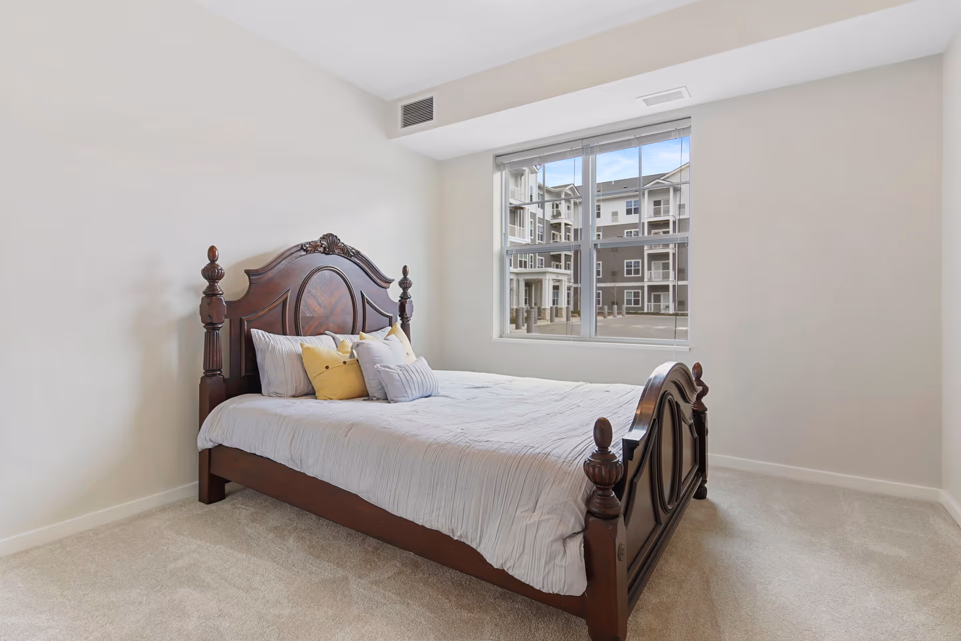 A bedroom with a large wooden bed frame featuring intricate carvings, a neatly made bed with white bedding and several pillows, a large window showing an exterior view of a multi-story building, and light-colored carpeted floor.