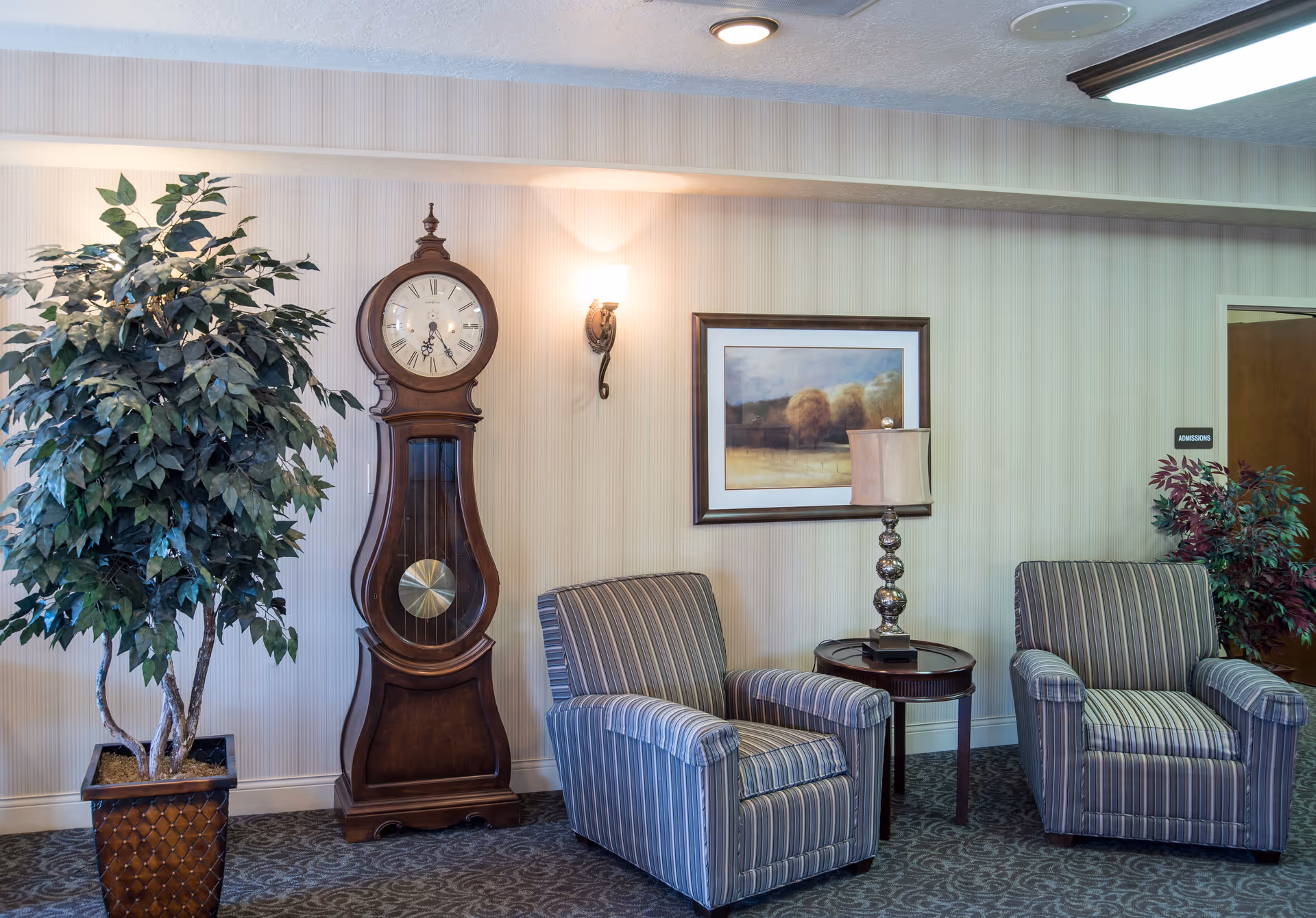 A cozy sitting area with two striped armchairs separated by a small round table with a decorative lamp. A large wooden grandfather clock stands against the wall next to a tall potted plant. A framed landscape painting hangs on the wall above the table. The wall has vertical striped wallpaper and a wall sconce light fixture is illuminated above the clock.