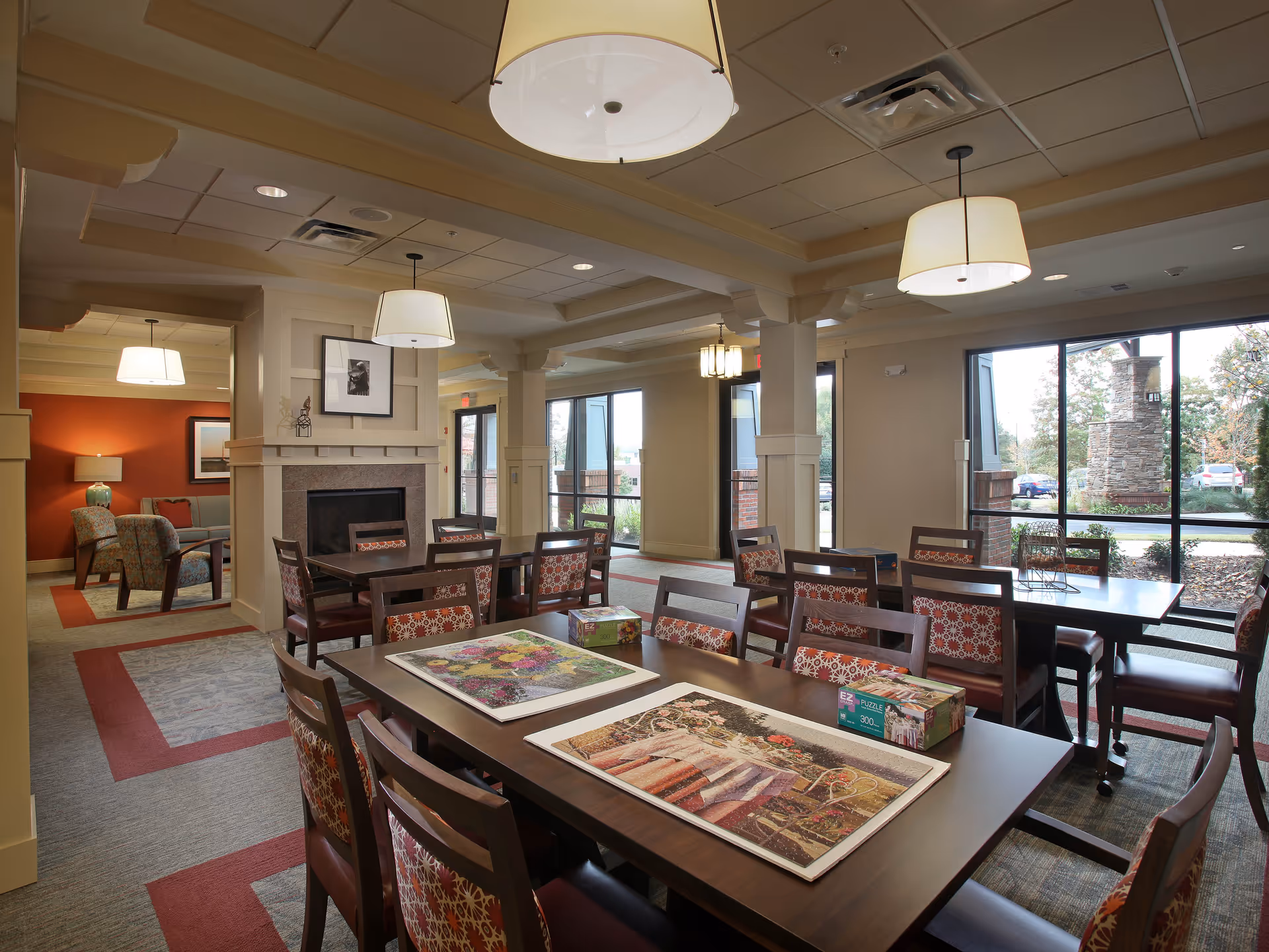 A well-lit common area in a senior living facility featuring multiple tables and chairs with patterned upholstery. Two tables in the foreground have partially completed jigsaw puzzles on them. The room has large windows allowing natural light to enter, a fireplace with framed artwork above it, and modern hanging light fixtures. In the background, there is a seating area with armchairs and a lamp against a red accent wall.
