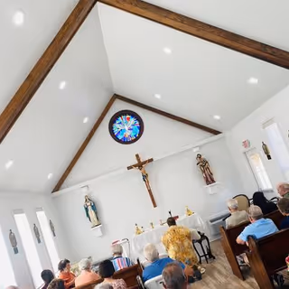 Interior of a small chapel with a high vaulted ceiling and wooden beams. A crucifix and religious statues are mounted on the front wall above an altar. Several elderly people are seated in pews facing the altar, participating in a religious service.