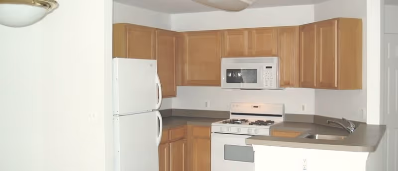 A kitchen area with wooden cabinets, a white refrigerator, a white microwave mounted above a white gas stove, and a countertop with a sink. The walls are white and the kitchen has a simple, clean design.