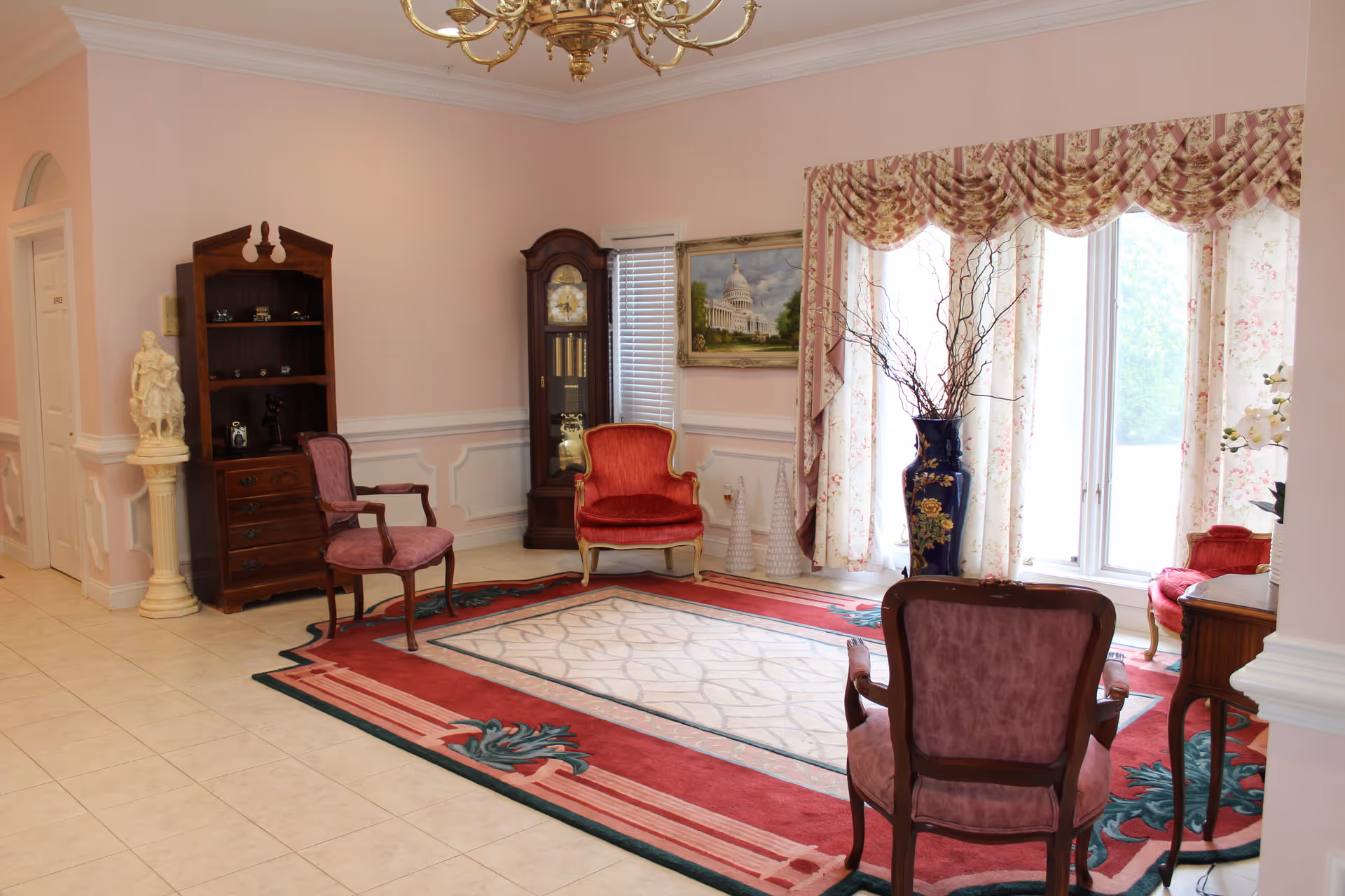 Sunlit formal sitting room with upholstered chairs, a grandfather clock, bookshelf, decorative rug and a large window with floral curtains.