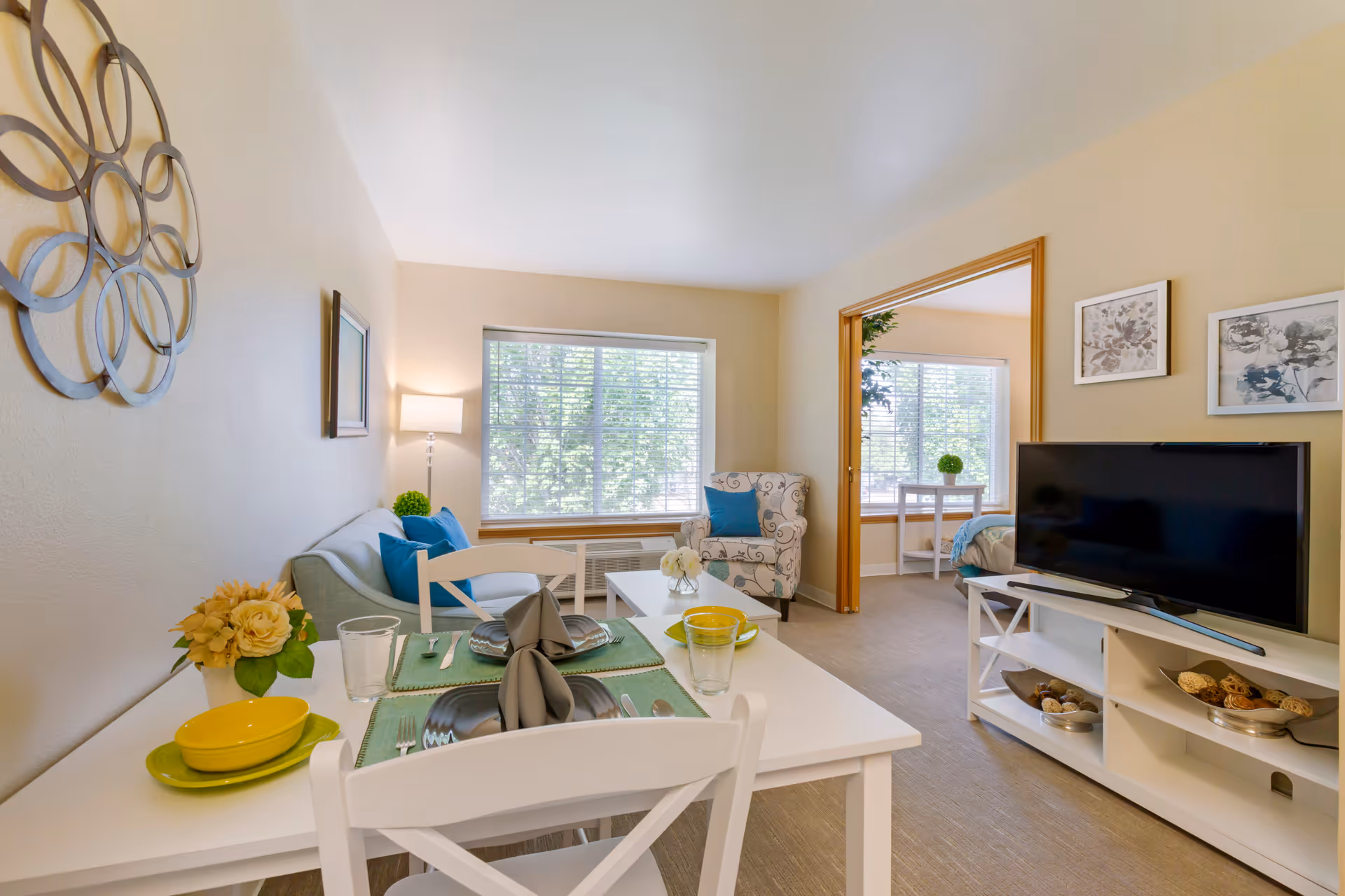 A bright and cozy living area in a senior living facility with a white dining table set for two in the foreground, a light blue sofa with blue cushions, a patterned armchair, a white coffee table, and a flat-screen TV on a white TV stand. Large windows with blinds allow natural light to fill the room. A bedroom with a bed and a small table with a plant is visible through an open doorway.