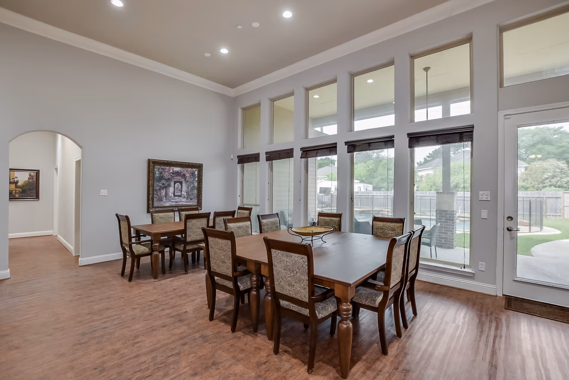 A spacious dining room with two wooden tables surrounded by upholstered chairs. The room features large windows with blinds, allowing natural light to enter and providing a view of an outdoor area with greenery and a patio. The walls are painted light gray, and there is a framed painting on one wall. The floor is wood, and the ceiling has recessed lighting.