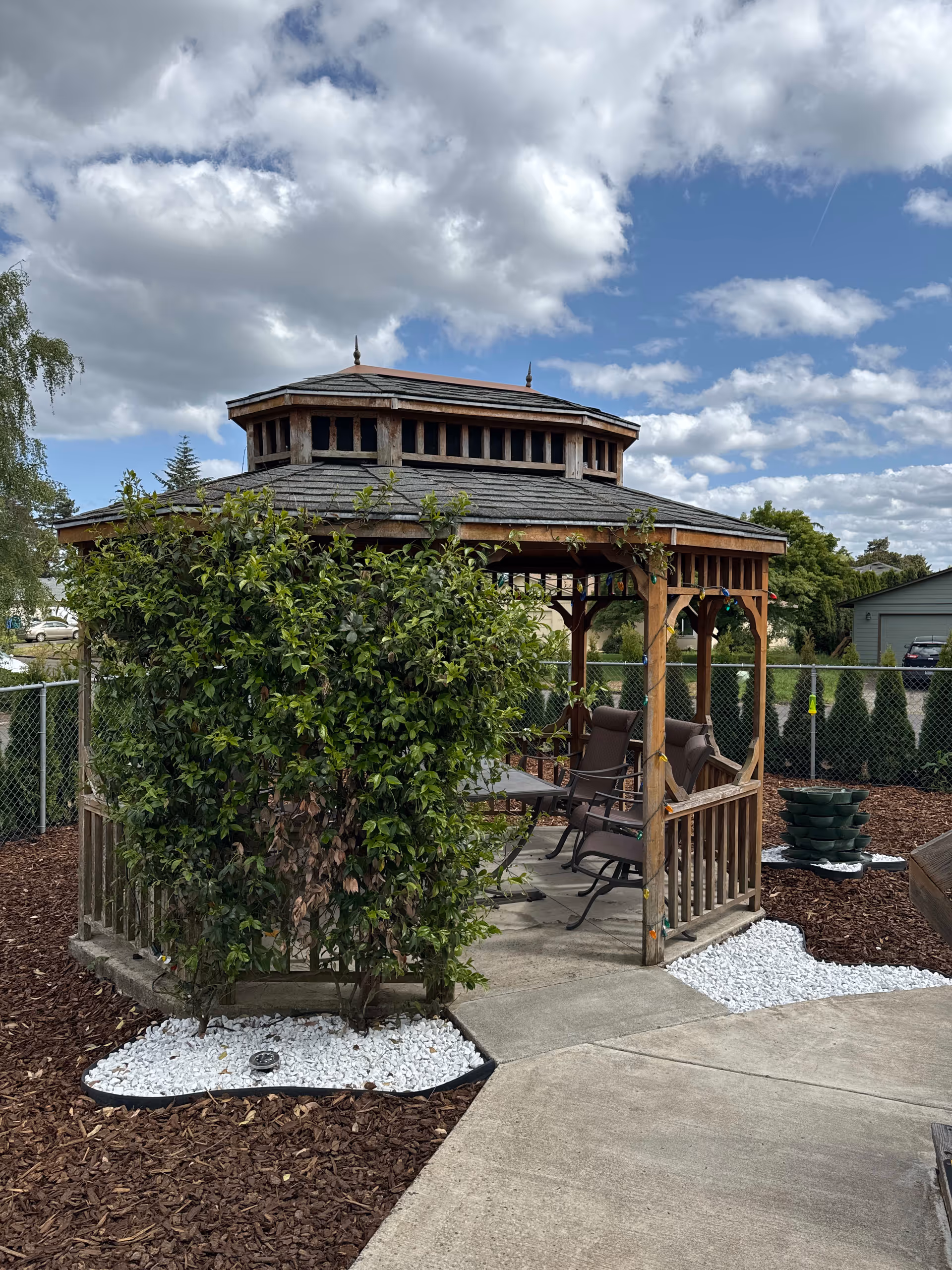 A wooden gazebo with a shingled roof in an outdoor garden area, surrounded by mulch and white decorative rocks. There are several cushioned chairs and a table inside the gazebo. The area is fenced with a chain-link fence and there are trees and shrubs around. The sky is partly cloudy.