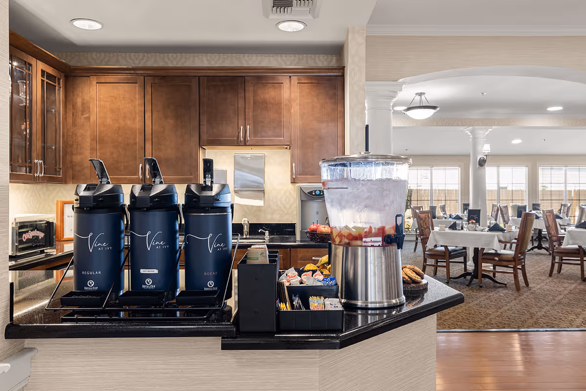 A coffee and beverage station with three large coffee dispensers labeled Vine at Ivy, a water dispenser with fruit-infused water, and a selection of condiments and snacks. In the background, there is a dining area with tables and chairs set for meals, featuring white tablecloths and napkins.