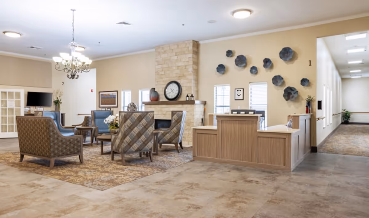 A spacious and well-lit common area in a senior living facility featuring a reception desk on the right, a stone fireplace with a clock above it in the center, and several upholstered armchairs arranged around a coffee table on a patterned rug. The walls are decorated with blue flower-shaped wall art and framed pictures. A chandelier hangs from the ceiling, and a hallway extends to the right.
