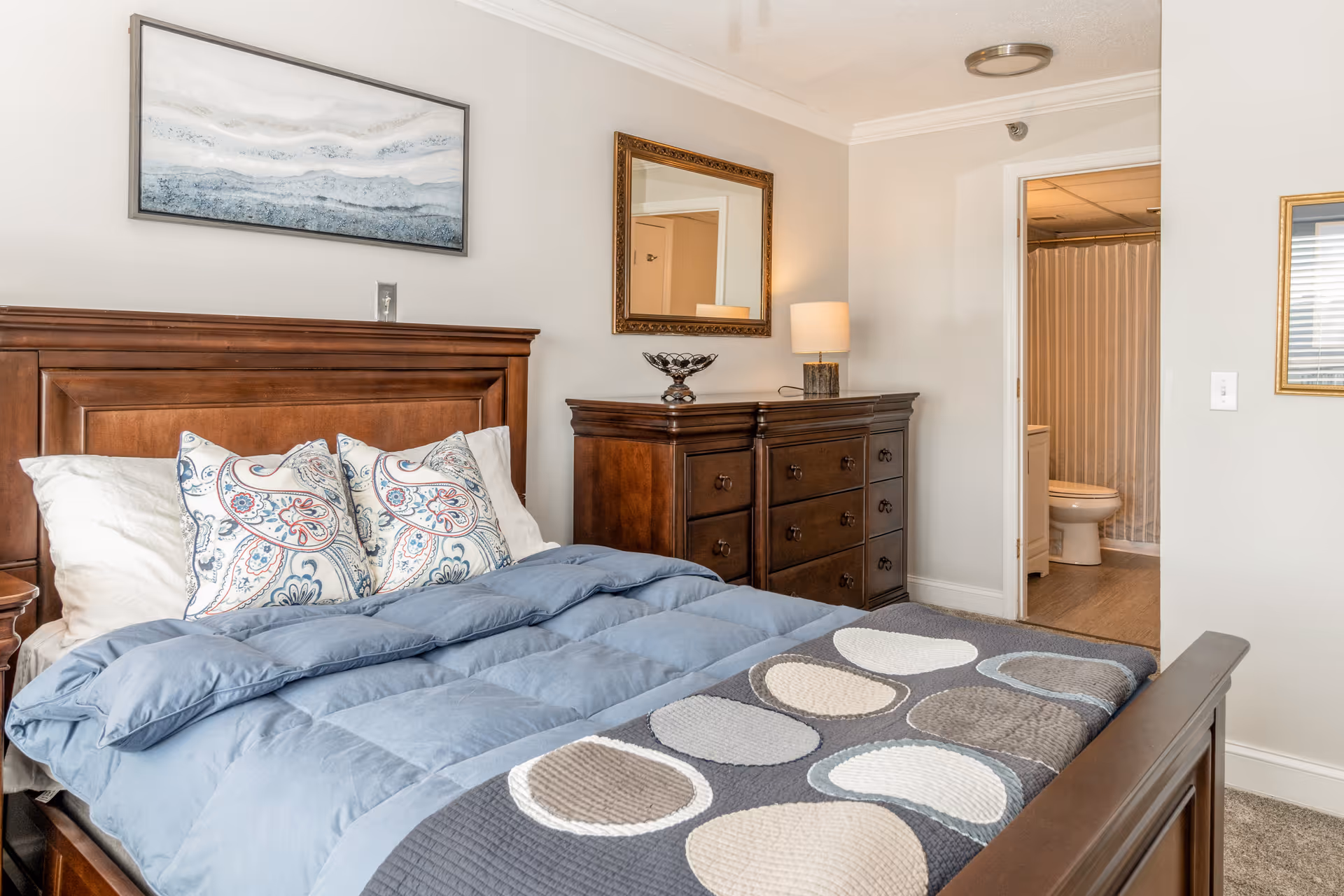 A bedroom with a wooden bed frame and headboard, a blue comforter, and three decorative pillows with a paisley pattern. There is a wooden dresser with a lamp and decorative bowl on top, a framed mirror above the dresser, and a framed abstract painting on the wall above the bed. An open door reveals a bathroom with a toilet and a shower with a striped curtain.