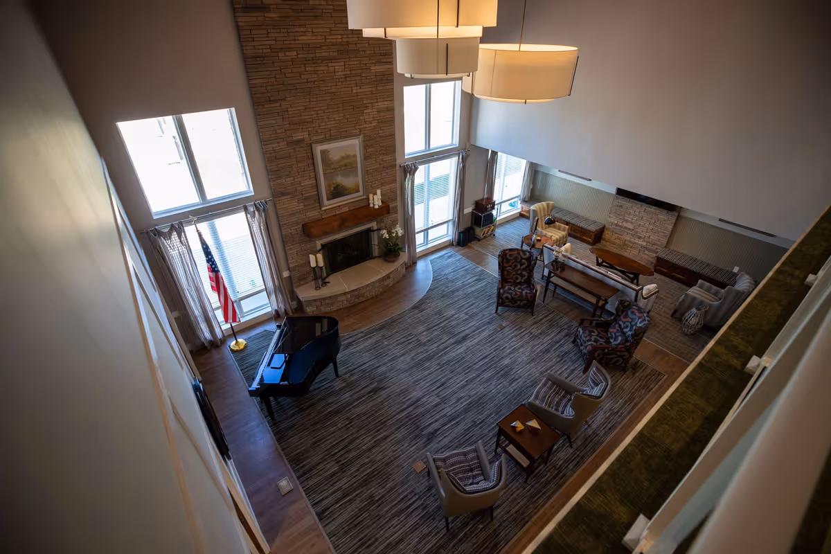 A spacious living room with a large area rug, multiple chairs arranged around a coffee table, a grand piano near a window with an American flag, and a stone fireplace with a painting above it. The room has high ceilings and large windows allowing natural light to enter.