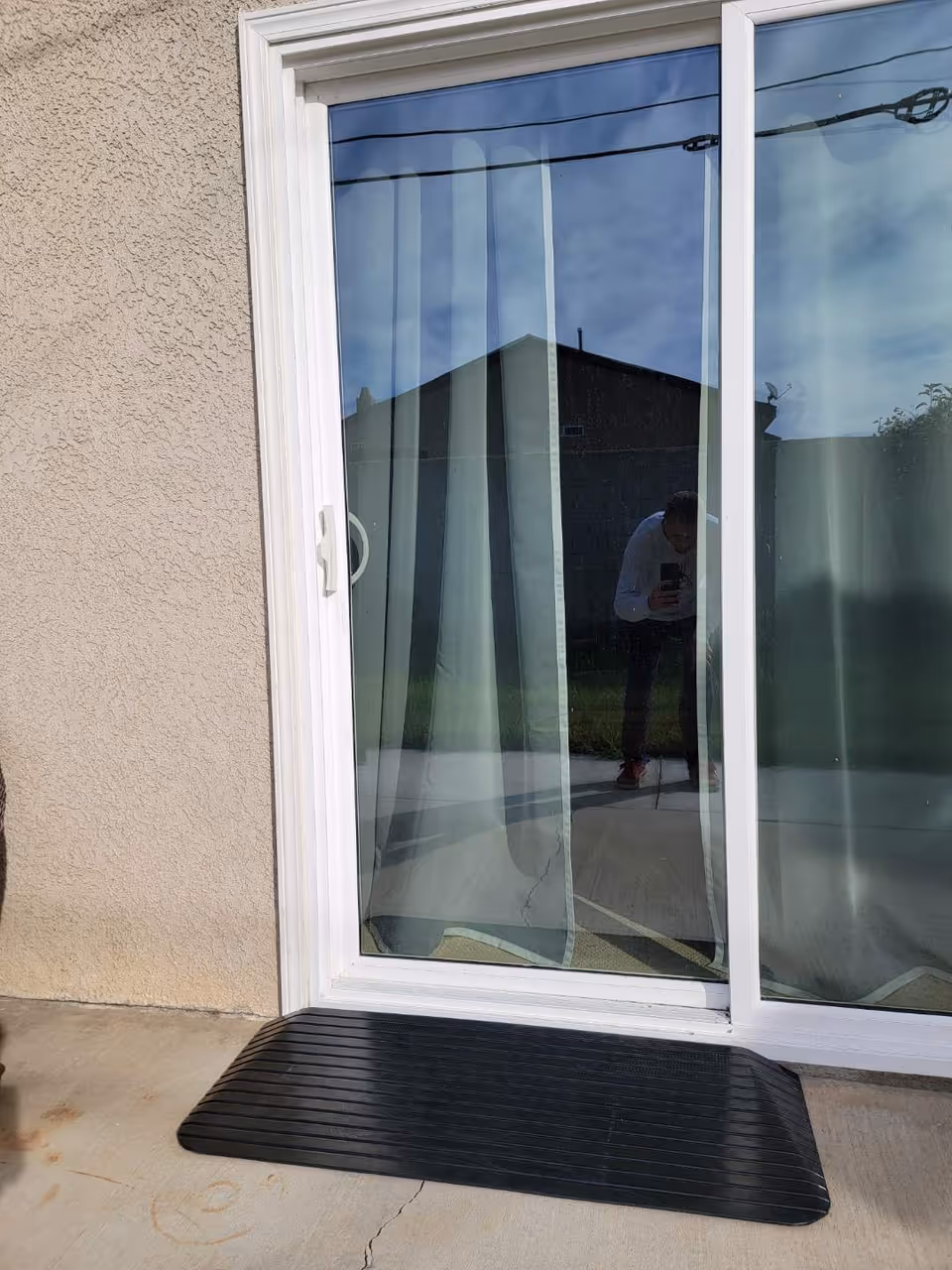 A sliding glass door with white frame and sheer curtains inside. The door reflects an outdoor scene including a building, power lines, and a person taking the photo. There is a black rubber ramp placed at the base of the door on a concrete surface.