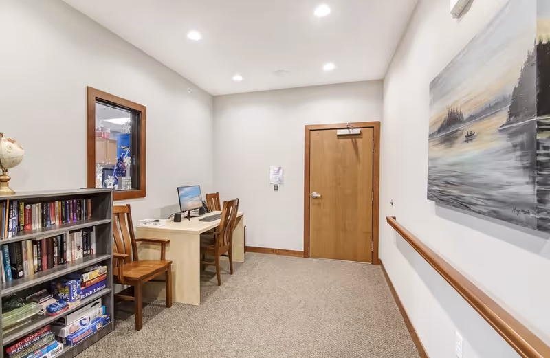 Small assisted-living common room with a desk, chairs, bookshelf and framed artwork on the wall.